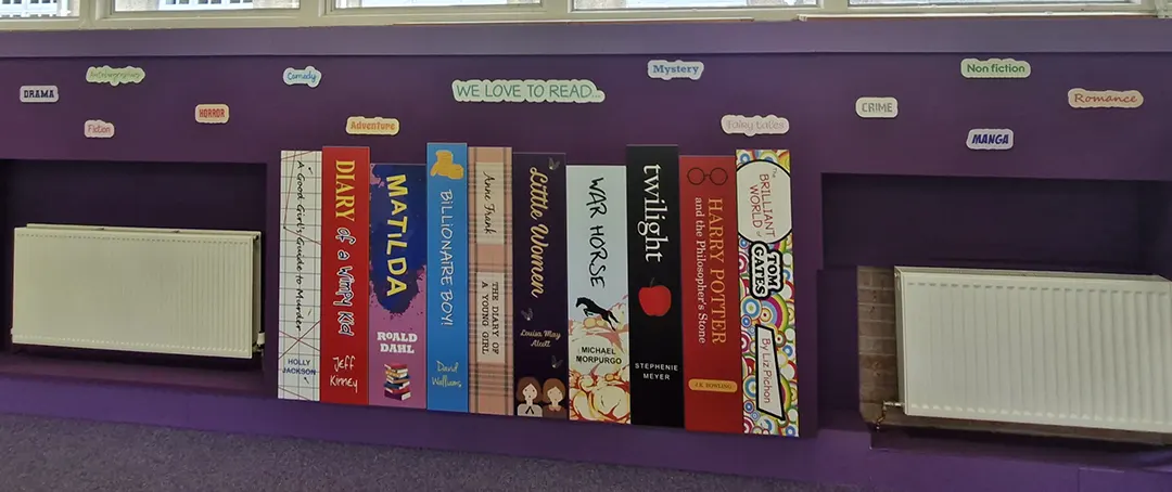 Book spine wall graphics on a school corridor wall featuring popular primary age book titles, with book genre words scattered across the wall above the book spines.